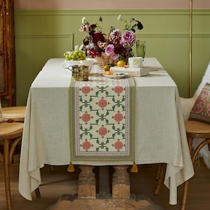 Puede incluir: Una mesa de comedor con un mantel crema y un camino de mesa floral. El camino de mesa presenta un patrón repetido de flores rosas y hojas verdes. Una cesta de flores, fruta y un candelabro están sobre la mesa. El ambiente es acogedor.