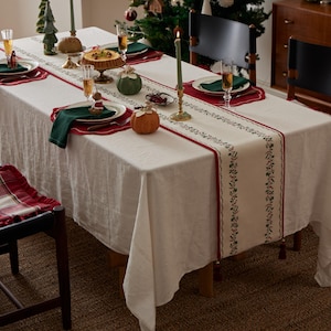 May include: A Christmas dinner table setting with a white tablecloth and a red and green runner. Plates, napkins, and decorative pumpkins are arranged on the table. Candles and a Christmas tree create a festive atmosphere. The table is set for a holiday meal.