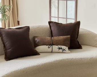 May include: A white, fluffy couch with three pillows. Two pillows are brown with dark brown stripes and one pillow is brown leather with a white pillow with a black bamboo design on top.