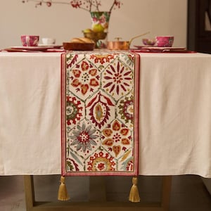 Può includere: Runner da tavolo rettangolare con sfondo color crema e motivo ripetuto di forme floreali e geometriche rosse, verdi e gialle. Il runner ha un bordo rosso e nappe dorate a ciascuna estremità. È posto su una tovaglia color crema.