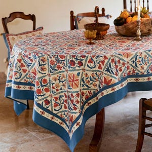 May include: A round tablecloth featuring a floral design in red, blue, and beige. The tablecloth has a blue border and is set on a wooden table. The table is set with a basket, bowl of fruit, and candle.