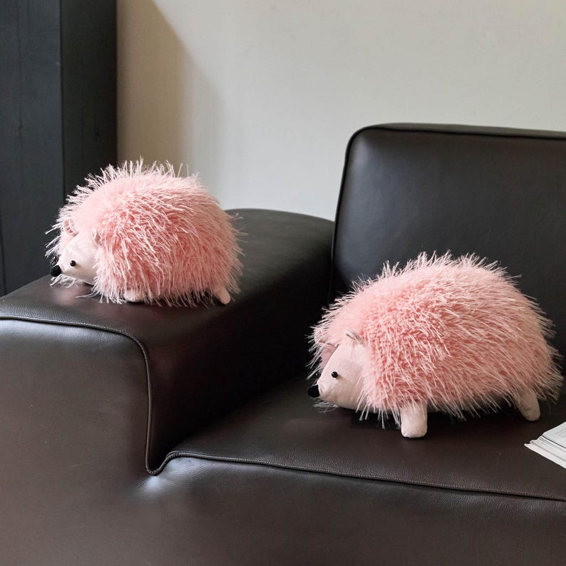 May include: Two plush pink hedgehog stuffed animals with soft, fluffy fur. The hedgehogs have white faces, small black eyes, and white feet. They are resting on a dark brown leather couch.