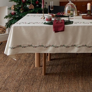 Op de afbeelding: Een crèmekleurig tafelkleed met een groene en rode botanische rand, op een houten tafel. De tafel is gedekt voor een feestelijke maaltijd, met een kerstboom op de achtergrond. Een mok met een kerstboomontwerp is zichtbaar.