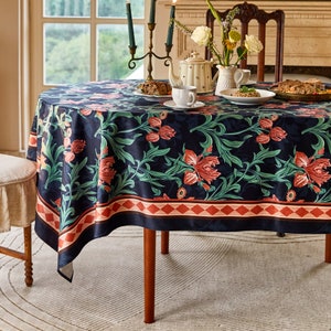 May include: A black and green floral tablecloth with a red and white border. The tablecloth is laid on a round wooden table with a vase of flowers and a teapot on top.