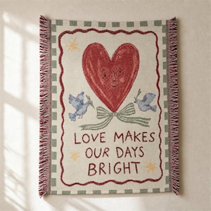 May include: A woven throw blanket featuring a central red heart with a face, flanked by two blue birds holding olive branches. Below, the text reads LOVE MAKES OUR DAYS BRIGHT. The design includes a green chequered border and fringed edges.
