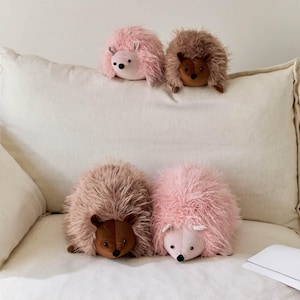 May include: Four plush hedgehog toys in shades of pink and brown rest on a white surface. The hedgehogs have soft, fluffy fur and contrasting brown or pink faces with black eyes and noses. The toys are arranged in two rows.
