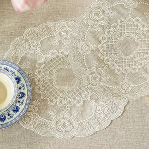 Puede incluir: Doilies de encaje blanco con intrincados patrones florales, superpuestos uno encima del otro. Los doilies están sobre un mantel de lino beige.