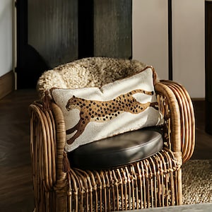 May include: A rectangular cushion featuring a cheetah design with brown spots on a cream background. The cushion rests on a dark brown leather seat of a wicker chair. Brown tassels adorn the corners.