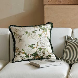 Pode incluir: Almofada quadrada com um design floral em branco, verde e castanho. A almofada tem uma borda verde escura com babados. Um livro com a palavra "PARIS" está no sofá. Outra almofada com listras pretas e brancas também está presente.