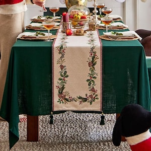 Può includere: Una tavola festosa apparecchiata per un pasto festivo. La tavola è coperta da una tovaglia verde scuro e da un runner color crema decorato con pigne, bacche e verde. La tavola è apparecchiata con piatti, bicchieri e un centrotavola.