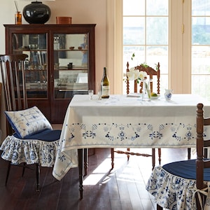May include: A dining room scene with a white tablecloth featuring a blue floral border, set with wine glasses and a bottle. Wooden chairs with blue cushions and matching patterned skirts surround the table. A cabinet and window are in the background.