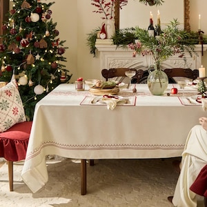 May include: A festive Christmas dining scene. A decorated tree with ornaments and lights stands near a table set with a cream-colored tablecloth with a red border. The table is set with plates, glasses, and a centerpiece.