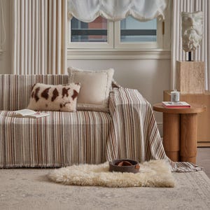 May include: A living room scene featuring a striped sofa with throw pillows and a matching throw blanket. A sheepskin rug and a small wooden side table with a candle and book are also present. The color palette includes beige, brown, and cream tones.