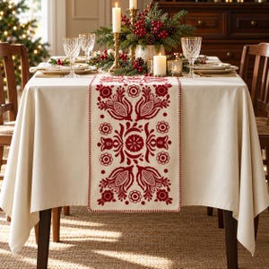 May include: A cream-colored tablecloth featuring a central runner with intricate red floral embroidery. The design includes symmetrical stylized flower motifs and circular patterns, finished with a stitched edge detail on the runner.