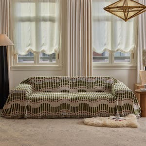 Könnte beinhalten: Ein Sofa, das mit einer gemusterten Decke bedeckt ist. Die Decke hat ein sich wiederholendes Design aus grünen vertikalen Linien und roten Punkten auf cremefarbenem Hintergrund. Das Sofa steht vor zwei Fenstern mit transparenten Vorhängen.