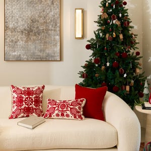 May include: A festive living room with a decorated Christmas tree, red cushions, and a white sofa. Two decorative cushions with red floral patterns and a plain red cushion are on the sofa. A framed artwork hangs on the wall.