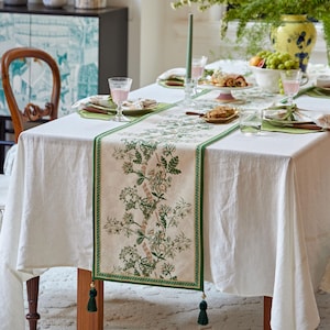 Può includere: Una tovaglia di lino bianca con un runner da tavolo floreale verde e beige. Il runner presenta un design botanico con finiture e nappe verdi. La tavola è apparecchiata con bicchieri, piatti e una candela.