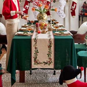 以下が含まれることがあります： 緑色のテーブルクロスに、白と緑の刺繍が施されたテーブルランナーが置かれ、クリスマスのモチーフが描かれています。テーブルにはグラスと皿が置かれています。