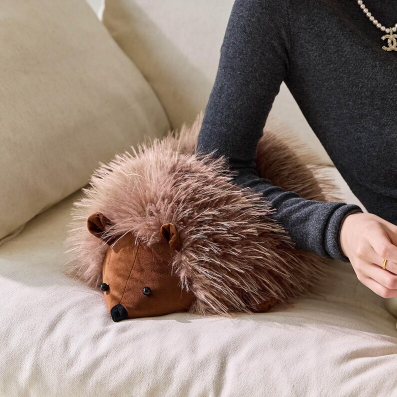 May include: A plush hedgehog pillow with a brown body and soft, light brown fur. The hedgehog has small, black eyes and a black nose. The pillow is resting on a cream-colored surface.