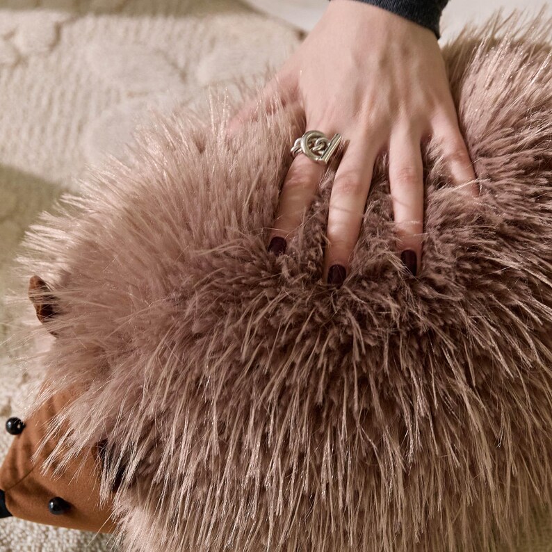 May include: A plush, brown hedgehog-shaped pillow with a soft, textured surface. The pillow has a brown snout and black bead eyes. A silver ring is on a hand resting on the pillow.