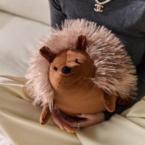 May include: A plush hedgehog toy with a brown body, ears, and legs, and a soft, light brown spiky exterior. The toy has small black eyes and a black nose. The hedgehog is a soft, cuddly toy.