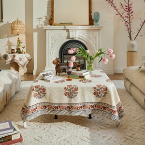 Puede incluir: Un mantel blanco con estampado floral y borde azul oscuro. El mantel está puesto sobre una mesa con una copa de vino tinto, una figurita de madera y una pila de libros. La mesa está en una habitación con una chimenea y un jarrón de flores rosas.