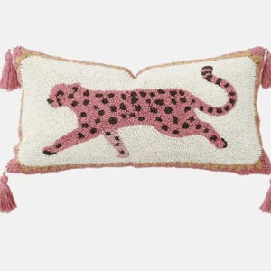 Puede incluir: Un coj&iacute;n rectangular con base de color crema y un dise&ntilde;o texturizado de hilo enganchado que muestra un leopardo rosa con manchas marr&oacute;n oscuro. El coj&iacute;n presenta un borde rosa y borlas rosas a juego en cada una de las cuatro esquinas.