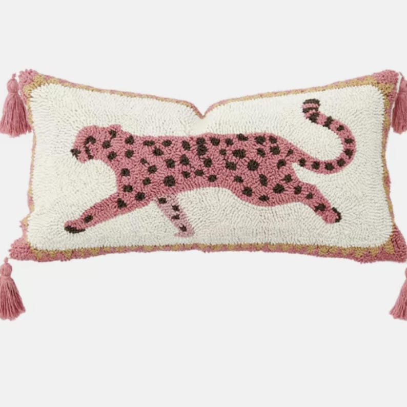Puede incluir: Un coj&iacute;n rectangular con base de color crema y un dise&ntilde;o texturizado de hilo enganchado que muestra un leopardo rosa con manchas marr&oacute;n oscuro. El coj&iacute;n presenta un borde rosa y borlas rosas a juego en cada una de las cuatro esquinas.