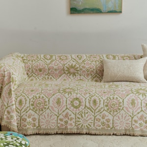 May include: A sofa draped with a floral patterned throw in pink, green, and cream. The throw has a fringed edge, and a matching cushion rests on the sofa. The pattern features stylized flowers and geometric shapes.