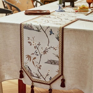 May include: A beige table runner with a brown border and tassels. The runner features a floral design with white blossoms and brown branches, and small blue birds.