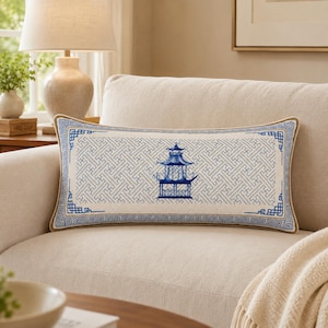May include: A rectangular lumbar pillow featuring a cream-colored fabric base with a central blue embroidered pagoda design. The pillow is accented by a geometric lattice pattern and a blue Greek key border, finished with a tan piped edge trim.