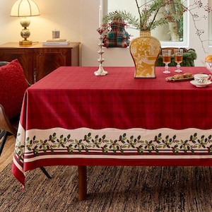 May include: A red tablecloth with a white border featuring a holly and berry design. The table is set with a vase, glasses, and a teacup, creating a festive holiday scene.