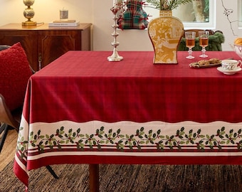 May include: A red tablecloth with a white border featuring a holly and berry design. The table is set with a vase, glasses, and a teacup, creating a festive holiday scene.