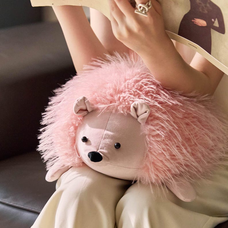 May include: A plush pink hedgehog toy with a soft, textured exterior. The toy has a light pink face, small ears, and black button eyes. The hedgehog is a cuddly, whimsical item.