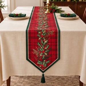 May include: A rectangular table runner featuring a red plaid pattern with a central botanical print of green holly leaves and red berries. The runner has a dark green border and a single dark green tassel at the pointed end.