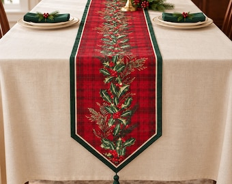 Pode incluir: Um caminho de mesa retangular com um padrão de xadrez vermelho e uma estampa botânica central de folhas de azevinho verdes e bagas vermelhas. O caminho tem uma borda verde-escura e uma borla verde-escura na extremidade pontiaguda.