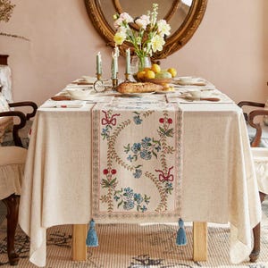 May include: A beige tablecloth with a floral runner featuring blue flowers, red cherries, and green leaves. The table is set with plates, silverware, candles, and a bread loaf. A vase of flowers and a mirror are in the background.
