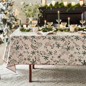 Puede incluir: Una mesa de comedor decorada para una celebración festiva. El mantel beige presenta un patrón de hojas verdes y bayas rojas. La mesa está adornada con copas, velas y arreglos florales, creando un ambiente cálido y acogedor.