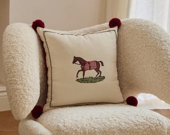 May include: Cream-colored square throw pillow with an embroidered horse design. The horse is brown with a red and white plaid blanket, standing on green grass. The pillow has a black stitched border and is adorned with red pom-poms.