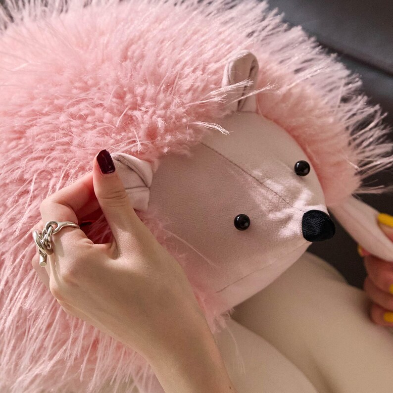 May include: A plush pink hedgehog stuffed animal with a soft, feathery texture. The hedgehog has a light pink face, ears, and body, with black eyes and a black nose. The toy is designed to be soft and cuddly.