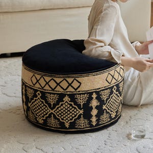 Puede incluir: Un otomán redondo negro con una banda estampada beige y negra alrededor de la base. El otomán tiene una apariencia suave y afelpada y está sobre una alfombra de color claro. Un vaso de agua está sobre la alfombra.