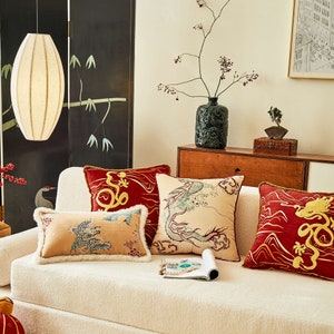 Può includere: Un divano bianco con quattro cuscini. I cuscini sono decorati con draghi ricamati e motivi floreali in tonalità di rosso, oro e blu. Il divano è davanti a uno schermo nero con un design di bambù.