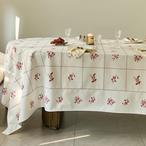May include: A cream-colored tablecloth with a floral pattern in red. The tablecloth is on a round table, set with glasses, a candle, and a plate of grapes. The floral design is repeated across the surface.