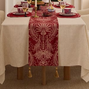 May include: A dining table set for a meal, with a cream tablecloth and a red table runner featuring a giraffe and palm tree design. The table is set with red placemats, plates, cups, and wine glasses. A gold candlestick and a small bowl are also present.