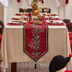 May include: A red and green plaid table runner with a holly and berry design. The runner has a green border and a tassel at the end. The runner is on a beige tablecloth with a Christmas-themed table setting.