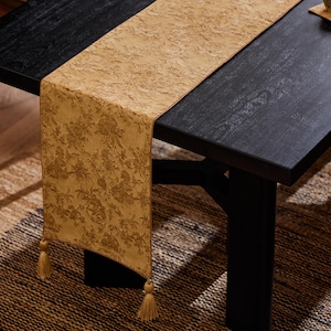 Puede incluir: Camino de mesa amarillo dorado con un estampado floral y borlas decorativas. El camino de mesa está colocado sobre una mesa de madera negra, agregando un toque de elegancia al comedor. Las borlas cuelgan en los extremos.