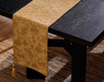 Op de afbeelding: Een goudgele tafelloper met een bloemenpatroon en decoratieve kwastjes. De loper ligt op een zwarte houten tafel en voegt een vleugje elegantie toe aan de eetkamer. De kwastjes hangen aan de uiteinden.
