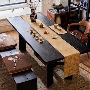 May include: A black wooden dining table set for tea. The table features a beige runner, small teacups, and a floral vase. Two benches with brown cushions are on the left. A wooden chair with a cushion is on the right.