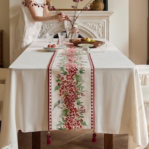 Op de afbeelding: Een rechthoekig crèmekleurig tafelkleed met een bloemen tafelloper. De loper heeft een rood bloemmotief met groene bladeren en een kleine vogel. De loper heeft een rode rand en kwastjes aan elk uiteinde. De tafel is gedekt met borden, glazen en een fruitschaal.