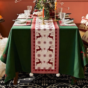 May include: A Christmas table setting with a green tablecloth and a red and white reindeer-themed table runner. The runner features reindeer and snowflake patterns. Place settings include plates, cutlery, and glasses. Candles and floral arrangements complete the festive scene.